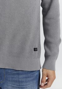 Grå strikkegenser med ribbede mansjetter og hem, med et lite svart merke på nedre venstre side. Brukt over blå denimjeans.