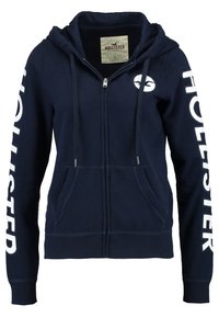Felpa con zip di colore blu navy realizzata in cotone, dotata di tasca frontale, lacci e la scritta "HOLLISTER" stampata in bianco su entrambe le maniche.
