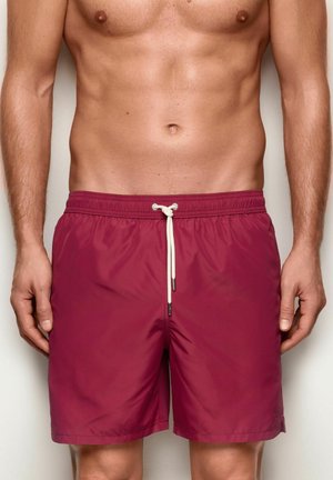 Torse masculino con bañador burdeos con cordón blanco, de pie frente a un fondo claro y liso.