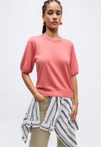 Nő fonott hajjal, arany fülbevalóval, korallszínű rövid ujjú pulóverben, bézs nadrágban és a derekára kötött csíkos ingben.