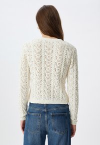 Strickpullover in Creme mit einem strukturierten offenen Spitzenmuster, langen Ärmeln und einem taillierten Saum, kombiniert mit hochgeschnittenen blauen Denim-Jeans.