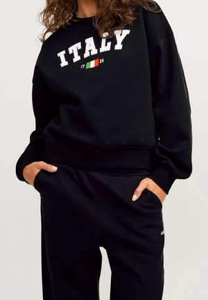 Personne portant un sweat-shirt noir avec "ITALY" et un drapeau italien, et un pantalon noir assorti, les mains dans les poches.