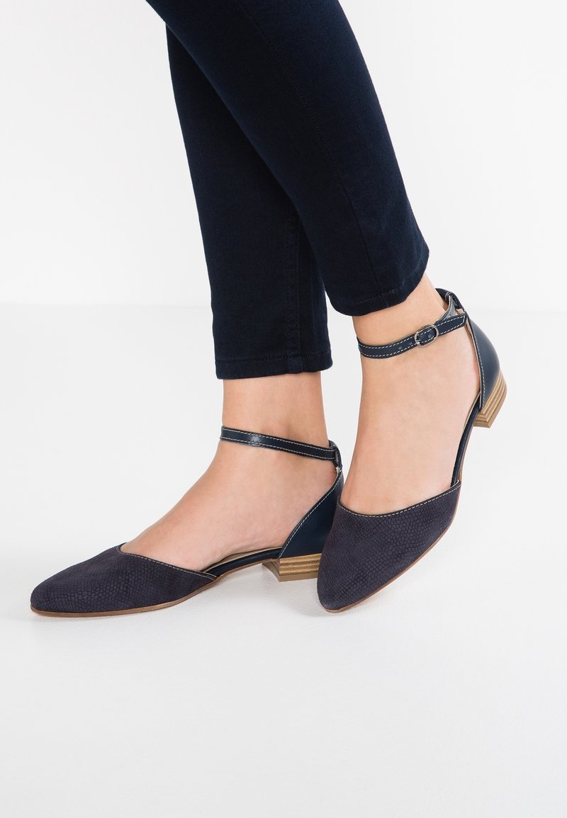 Chaussures plates bleu marine à bride à la cheville avec petits talons en bois, portées avec un pantalon ajusté foncé sur fond blanc.