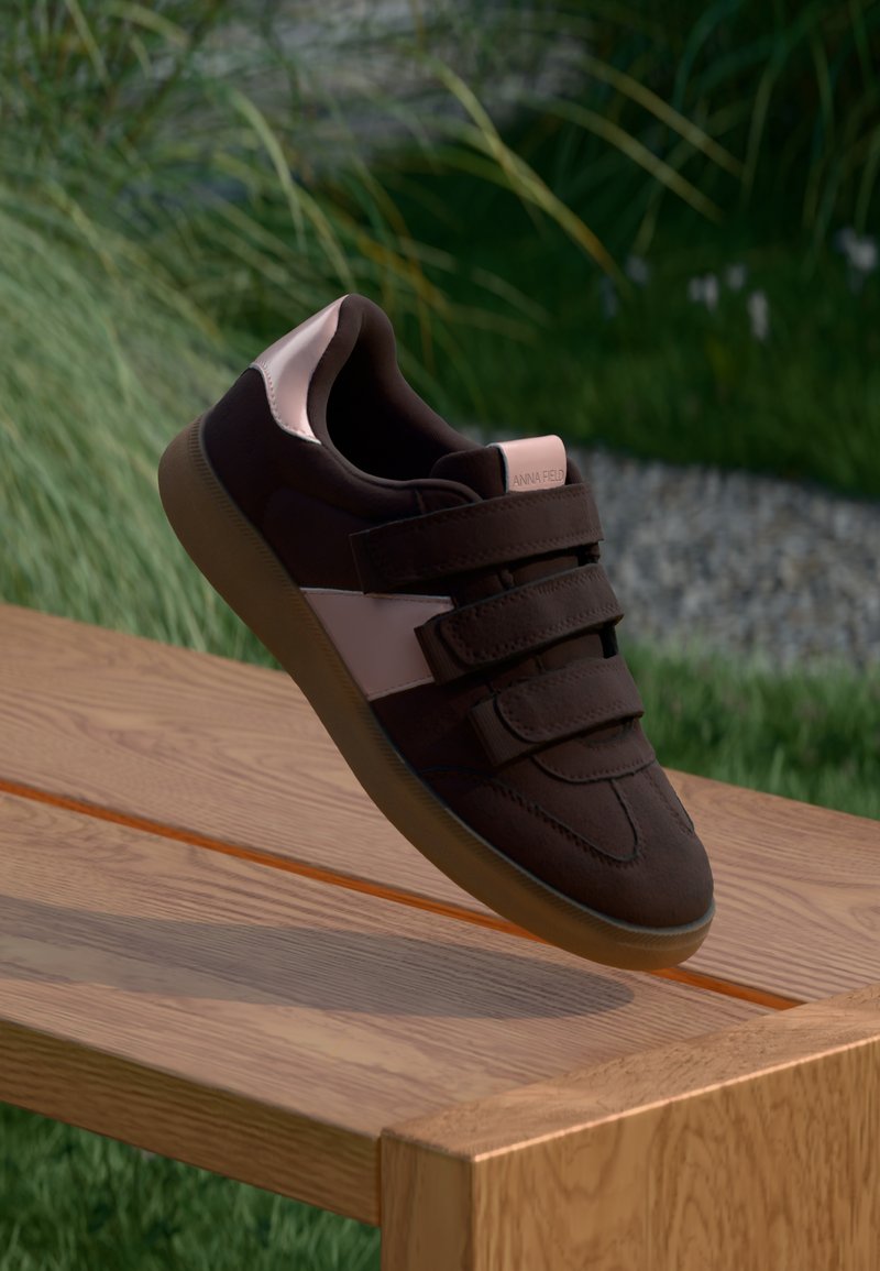 Baskets en daim marron avec une bande d'accent rose, comportant trois bandes Velcro et une semelle en caoutchouc texturé, posées sur une surface en bois.
