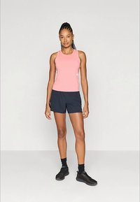 Moteris stovi vilkėdama rožinį be rankovių marškinėlį, juodus sportinius šortus, juodas kojines ir juodus sportbačius prieš vienodą šviesią foną.