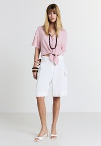 Donna in piedi che indossa una blusa leggera rosa chiaro a maniche corte con nodo, pantaloni corti bianchi alla ginocchia con tasche, sandali bianchi con tacco e braccialetti marroni.