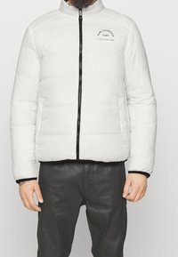 Vit puffjacka med hög krage, svart dragkedja och logotyp på vänster bröst. Har sömmar i paneler och svarta detaljer vid ärmsluten och nederkant.