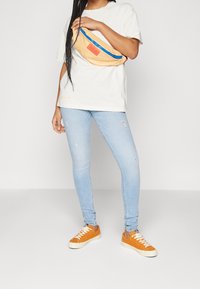 Mujer vistiendo jeans ajustados rotos de color azul claro, zapatillas naranjas, camiseta blanca de gran tamaño, y sosteniendo una riñonera beige con cremallera azul cruzada sobre el pecho.