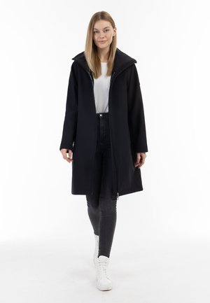 Junge Frau geht selbstbewusst, trägt einen langen schwarzen Mantel, weißes Hemd, dunkle Jeans und weiße Schnürstiefel vor weißem Hintergrund.
