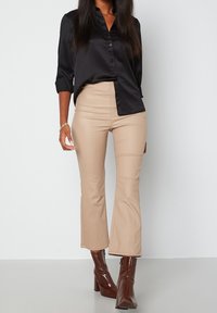 Chemise noire en satin à boutons avec des manches retroussées, associée à un pantalon beige à taille haute et évasé. Le modèle porte des bottes à talons brillantes de couleur marron.