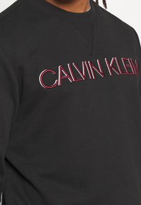 Felpa nera in cotone con logo ricamato "CALVIN KLEIN" rosso e bianco sul davanti, con collo a giro e tessuto testurizzato.