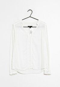 Camicia bianca a maniche lunghe realizzata in tessuto morbido, con scollo a V, chiusura a bottoni e dettagli plissettati vicino alle spalle. Appesa a un gancio nero.