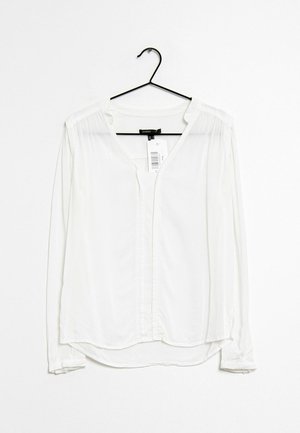Camicia bianca a maniche lunghe realizzata in tessuto morbido, con scollo a V, chiusura a bottoni e dettagli plissettati vicino alle spalle. Appesa a un gancio nero.