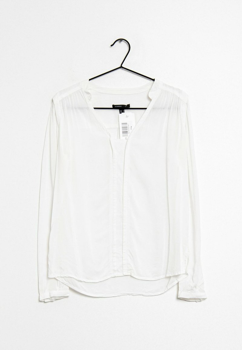 Camicia bianca a maniche lunghe realizzata in tessuto morbido, con scollo a V, chiusura a bottoni e dettagli plissettati vicino alle spalle. Appesa a un gancio nero.