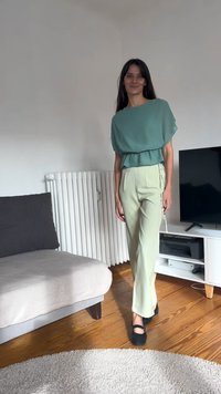 Voľná krátkym rukávom tyrkysová blúzka s volánovým lemom a svetlozelené vysoký pás nohavice. Čierne ploché topánky. Neutrálne vnútorné prostredie.