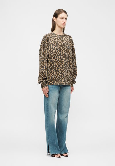 Ženska, oblečena v širok pulover z leopardjim vzorcem, široke bledomodre kavbojke z razporki spredaj in črne odprte pete, na belem ozadju.