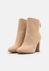 Bottes cheville en daim beige avec un bout pointu, un talon bloc et une languette de tirage à l'arrière. Texture lisse avec un léger éclat.