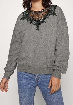 Femme portant un sweat-shirt gris à manches longues avec un détail en dentelle noire au niveau du col et un jean bleu, debout avec les bras détendus.