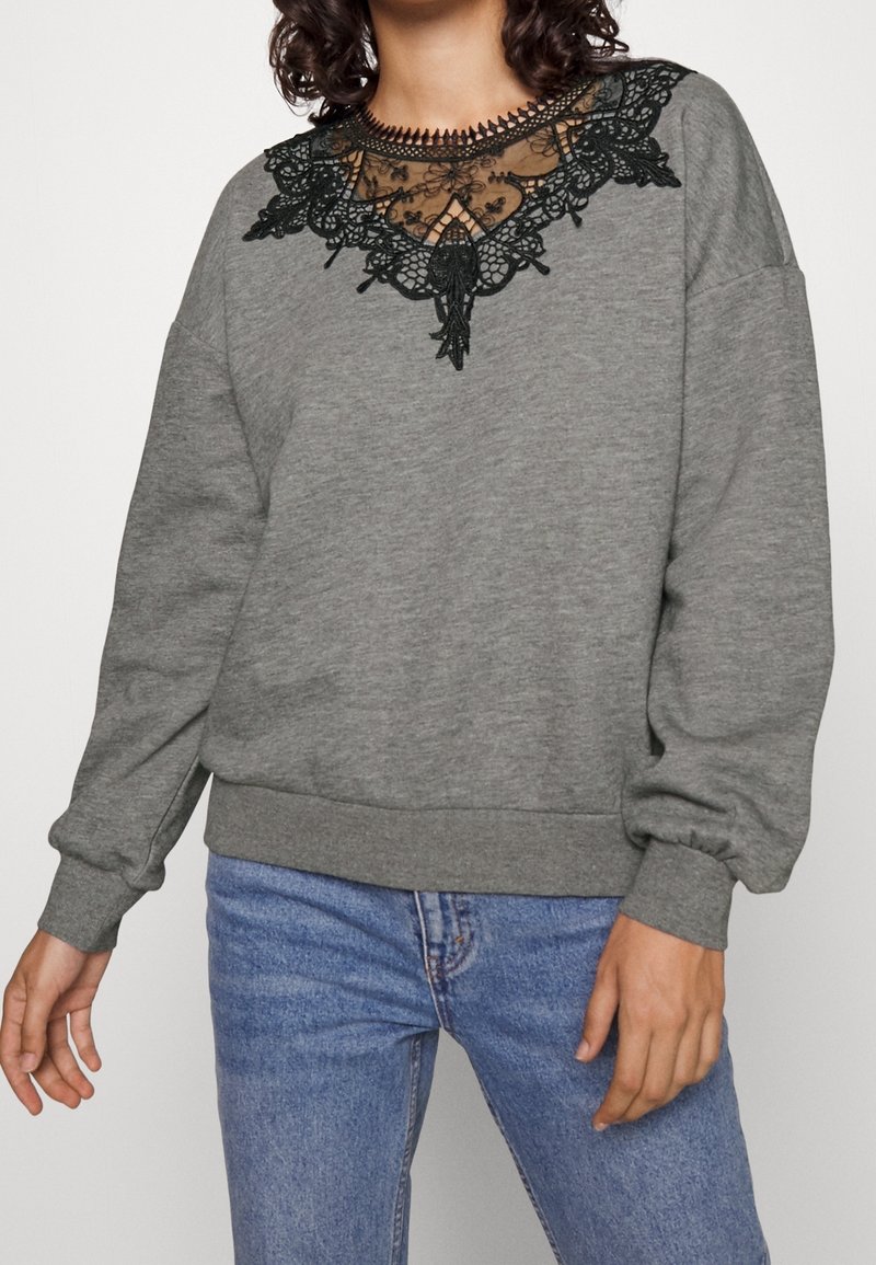 Femme portant un sweat-shirt gris à manches longues avec un détail en dentelle noire au niveau du col et un jean bleu, debout avec les bras détendus.