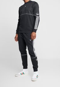 Hombre vestido con un chándal negro de Adidas con rayas y logotipos blancos, combinado con zapatillas blancas que tienen rayas verdes y rojas.