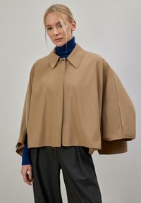 Veste beige à col avec fermeture à boutons, dotée de manches larges et d'un design élégant. Portée sur un col roulé bleu et un pantalon gris à carreaux.
