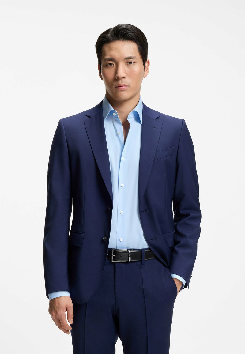 Homme debout face à l’avant, vêtu d’un costume bleu, d’une chemise bleu clair et d’une ceinture noire, une main dans la poche, sur fond uni.