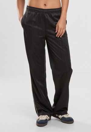 Osoba w luźnych czarnych spodniach dresowych z bocznymi pasami i niebiesko-białych trampkach, stojąca z rękami w kieszeniach.