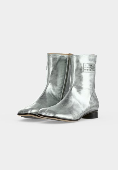Bottines argentées brillantes avec fermetures éclair latérales, talons bas noirs et une petite étiquette blanche avec des chiffres sur le côté extérieur de chaque botte.