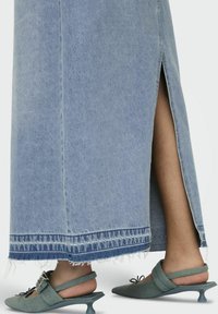 Jupe en denim bleu clair avec un ourlet effiloché et une fente latérale, portée avec des chaussures à talons décrochés vertes ornées d'un nœud et d'un talon incurvé.