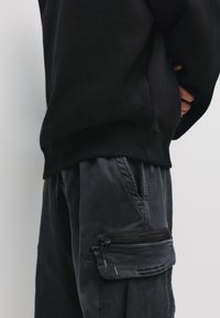 Hanorac negru cu tiv țesut, asortat cu pantaloni cargo închiși la culoare, cu un buzunar lateral cu fermoar și un material neted, texturat.