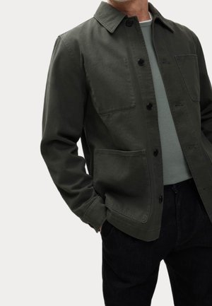 Mann trägt dunkelgrüne Knopfjacke mit großen Fronttaschen über einem hellgrauen Hemd und schwarzen Hosen, Hände in den Taschen.