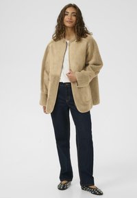 Naine, kes kannab helepruuni karusnahktagi, valget särki, tumedaid siniseid teksaseid ja musti naelu täis kingi, seisab ühevärvilise tausta ees.