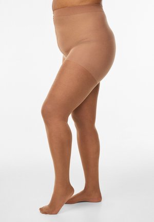 Stranski pogled na osebo, ki nosi prosojne hlačne nogavice v barvi kože, stoji bosa ob enostavni beli ozadju.