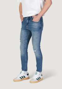 Plave skinny traperice s izblijedjelim dijelovima, laganim habanjem na koljenima i klasičnim dizajnom s pet džepova, nosene s bijelim i zelenim tenisicama.