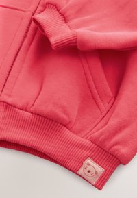 Koralrød sweatshirt med ribstrikkede ærmeafslutninger og kant. Har et lille bjørnepatch-label. Blød tekstur, afslappet design og forlomme.