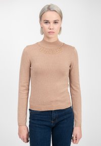 Una mujer con cabello corto rubio que lleva un suéter beis brillante con un escote de cuentas y jeans azul oscuro, de pie frente a un fondo blanco.