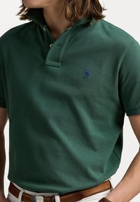 Grønn polo skjorte i teksturert stoff; har en to-knappers placket, hevet krage og liten blå logo på venstre bryst. Bært med et brunt belte.