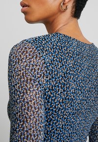 Vue de profil d’une personne portant un haut à motif floral bleu, noir et blanc avec une boucle d’oreille en anneau dorée, sur un fond uni.