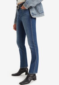 Jeans in denim blu scuro con vestibilità slim, caratterizzati da dettagli in borchie metalliche lungo le cuciture laterali, abbinati a stivaletti neri con tacco a blocco.