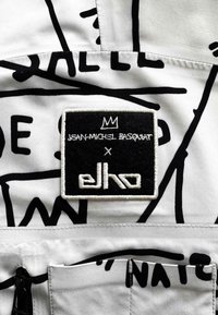 Biały materiał z czarnymi abstrakcyjnymi liniami; zawiera prostokątną naszywkę z czarnego filcu, białym tekstem i ikoną korony, oznakowaną marką "Jean-Michel Basquiat x elho".