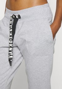 Šedé teplákové kalhoty s tenkým páskem a černo-bílým logem. Měkká látka, boční kapsy a pohodlný střih.
