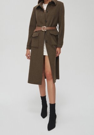 Manteau en laine marron avec col, fermeture à boutons, poches avant, et ceinture marron avec une boucle carrée. Porté par-dessus une robe blanche et des bottes noires.