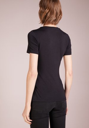 Personne aux cheveux bruns courts portant un t-shirt noir uni à manches courtes et un pantalon noir, vue de dos sur un fond neutre.