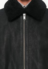 Black leather jacket with a textured finish, featuring a plush black collar, silver zipper, and gray checkered shirt underneath.