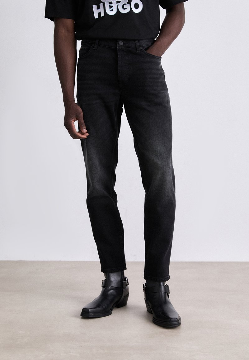 Jeans en denim noir à coupe droite, avec cinq poches et une décoloration subtile. Portés avec des bottines noires et un t-shirt noir à motif.