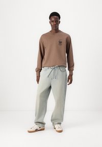 Felpa marrone con logo e maniche lunghe, abbinata a jeans di denim azzurro chiaro e dal taglio morbido. Le scarpe sono di un color avorio con dettagli scuri.