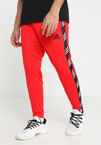 Homme portant un pantalon de survêtement rouge Adidas avec des rayures latérales noires et blanches et des chaussures de sport blanches avec des lacets noirs.