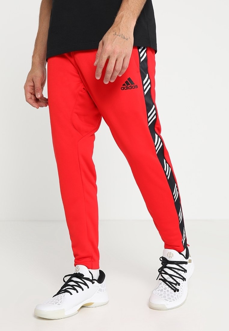 Homme portant un pantalon de survêtement rouge Adidas avec des rayures latérales noires et blanches et des chaussures de sport blanches avec des lacets noirs.