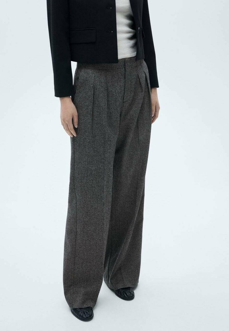 Femme porte un pantalon large en laine grise taille haute, un blazer noir court, un haut blanc et des chaussures noires, debout devant un fond clair uni.