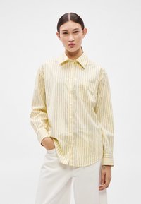 Dzeltena svītraina krekli ar pogām, apkakli, garām piedurknēm un krūšu kabatu. Uz gaiša fona ir vertikālas zilas svītras.